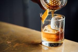 copo de whisky com gelo com rodela de laranja sobre mesa e uma coqueteleira