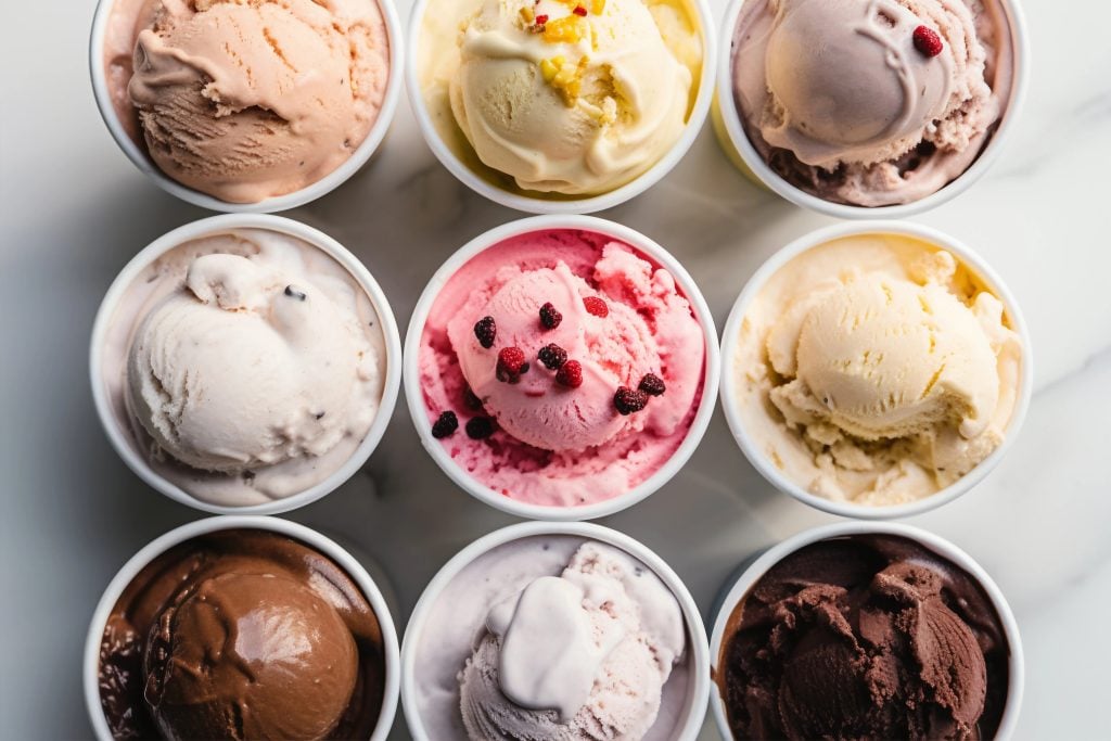 Vista superior de nove copos de sorvete colorido dispostos em uma grade, com sabores variados como chocolate, baunilha, frutas vermelhas, creme e outros, sobre uma superfície de mármore branco.